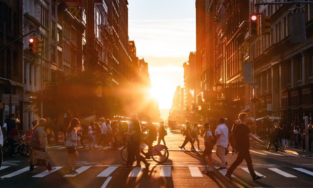 People walking through the busy intersection at 5th Avenue and 23rd Street in New York City on a summer day with sunset flare behind the background buildings and air pollution in the sky