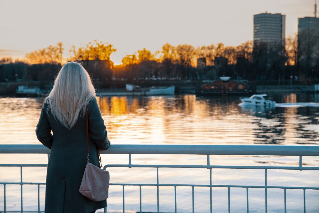 Rear view of white hair Woman enjoying the sunset over the city river. Female standing on the bridge enjoying cityscape at evening
