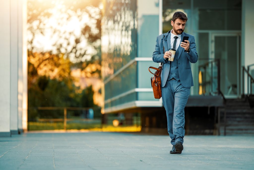 A businessman in a suit walks outside while checking his smartphone. He carries a coffee cup and a leather bag, enjoying a sunny day. 